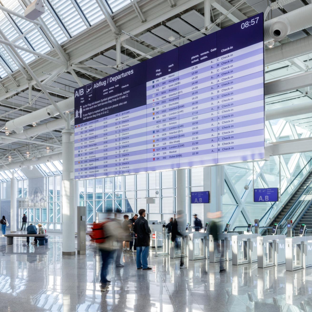 Lichtdurchflutete Abflughalle des Flughafens München mit Glasdach und Glasfassade. Unter einer großen digitalen Abfluganzeigetafel gehen Reisende mit Gepäck durch einen Self-Service-Bereich mit Check-in- und Bag-Drop-Automaten.