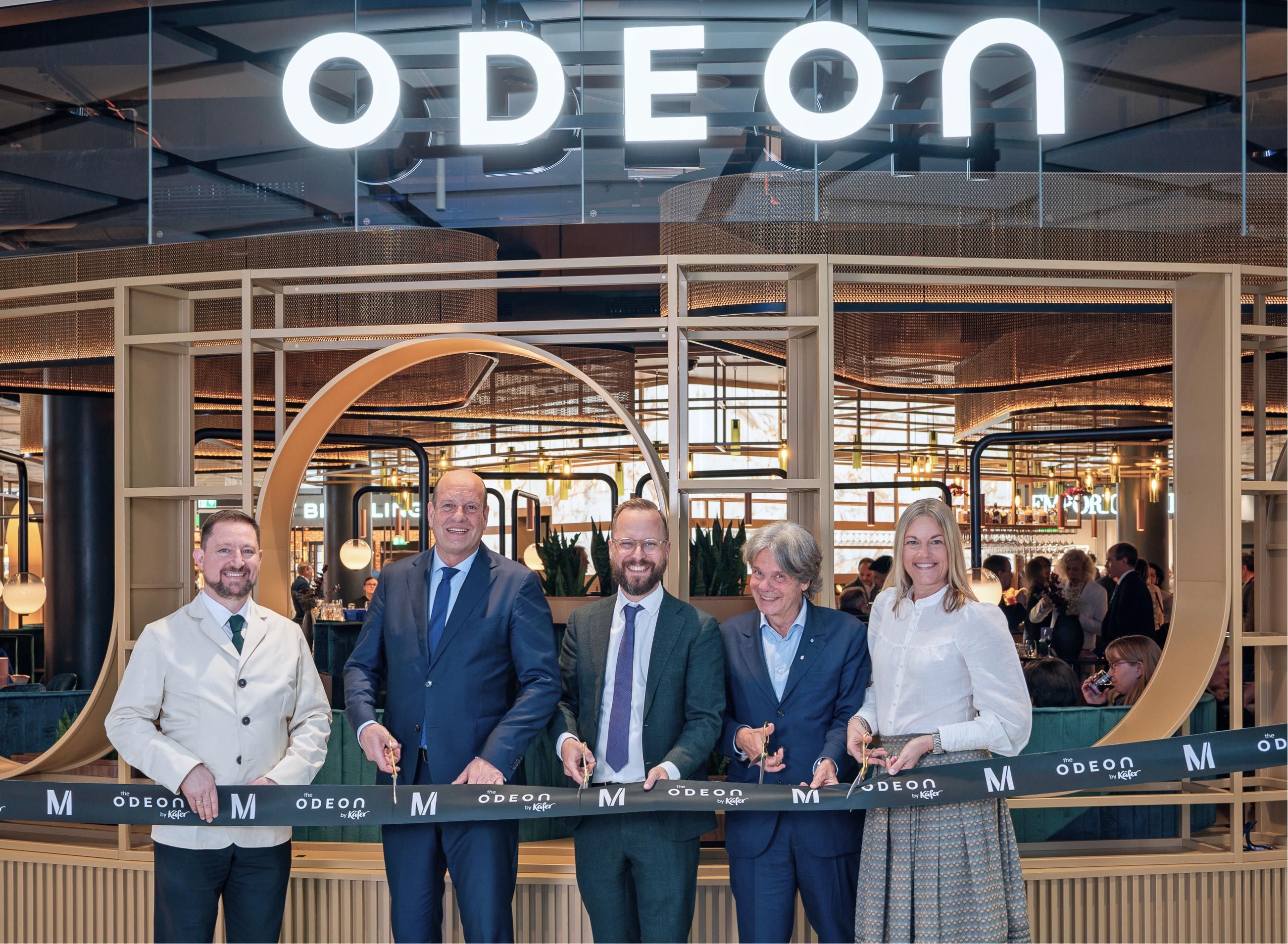 Feierliche Eröffnung des Gastronomiebereichs „the Odeon by Käfer“ am Flughafen München: Fünf Personen durchschneiden gemeinsam ein Eröffnungsband vor dem beleuchteten Odeon-Schriftzug, darunter Dr. Jan-Henrik Andersson, Geschäftsführer Commercial und Security.