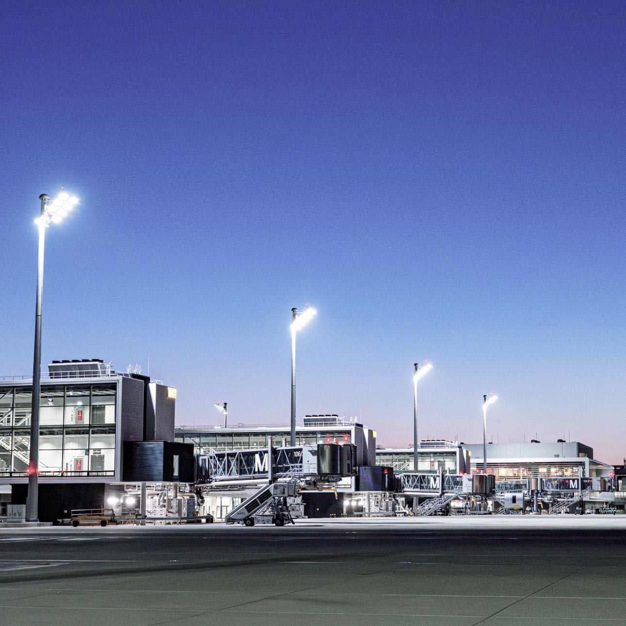 Terminalgebäude des Flughafens München mit Fluggastbrücken am Vorfeld in der Dämmerung, beleuchtet von hohen Flutlichtmasten vor tiefblauem Abendhimmel.