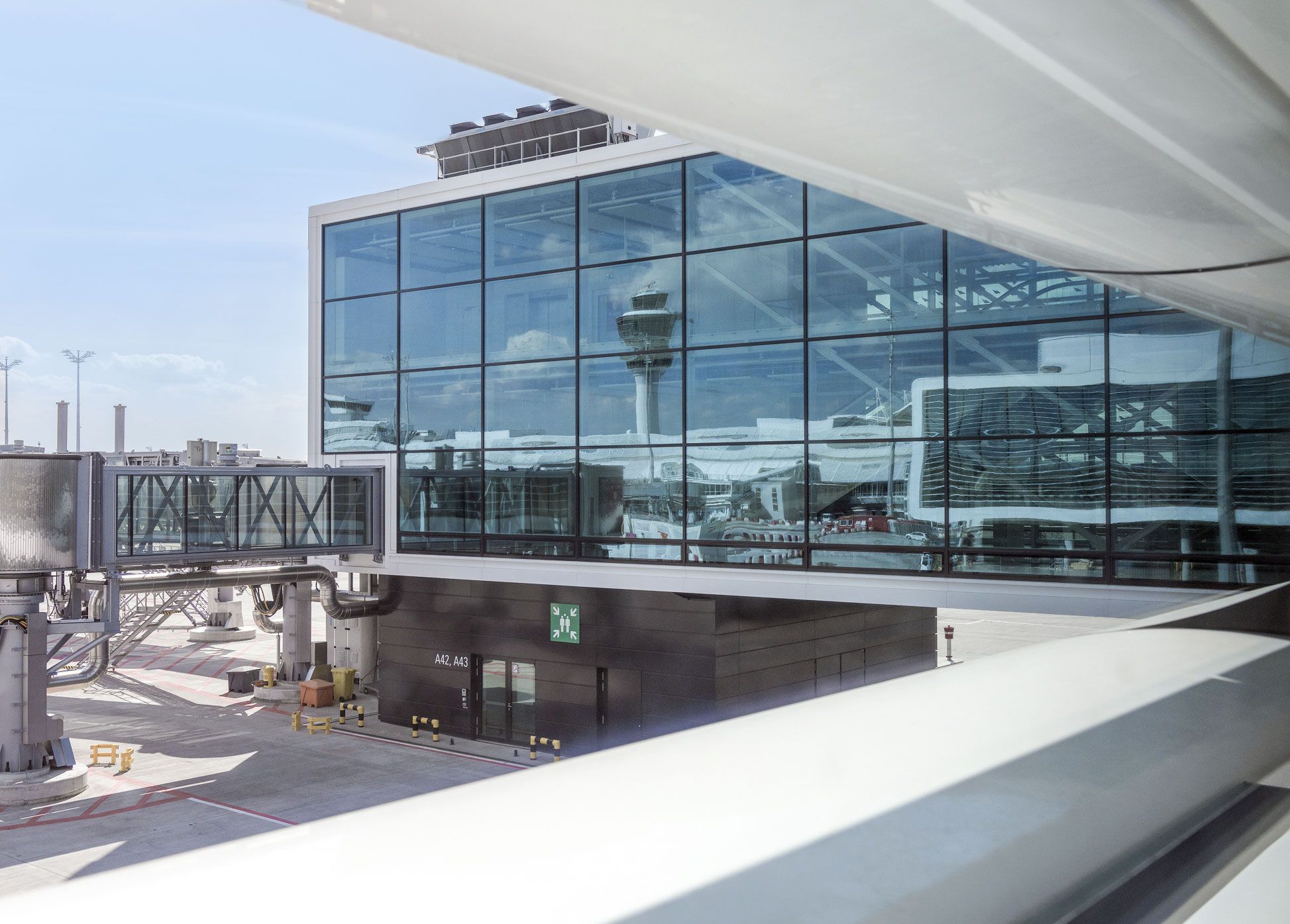 Verglaste Fassade eines Terminalgebäudes am Flughafen München mit angedockter Fluggastbrücke zu den Gates A42 und A43; in der Glasfront spiegelt sich der Tower.