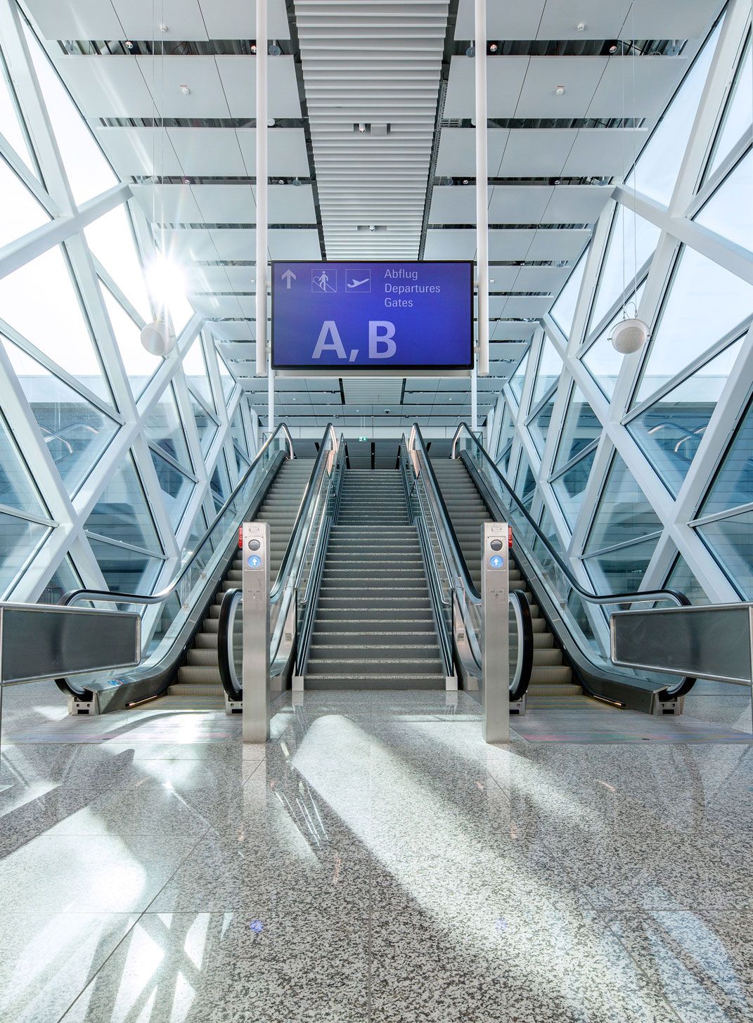 Rolltreppen in einer lichtdurchfluteten, verglasten Terminalhalle des Flughafens München, darüber ein Wegweiser „Abflug / Departures / Gates A, B“.