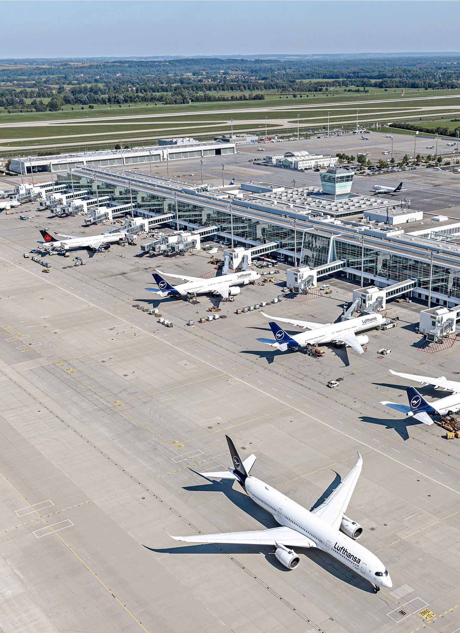 Luftaufnahme des Flughafens München mit Terminal, Vorfeld und Tower; mehrere Passagierflugzeuge stehen an den Fluggastbrücken, ein Lufthansa-Flugzeug rollt über das Vorfeld.