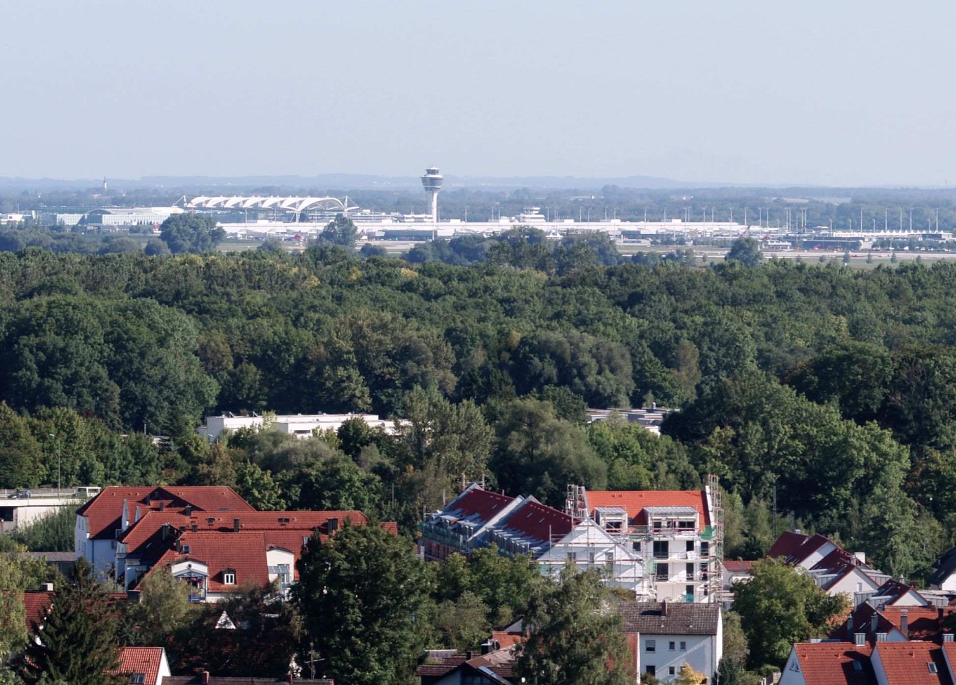 Blick über eine bewaldete Ortschaft mit Wohnhäusern auf den am Horizont liegenden Flughafen München mit Tower und Terminaldach.